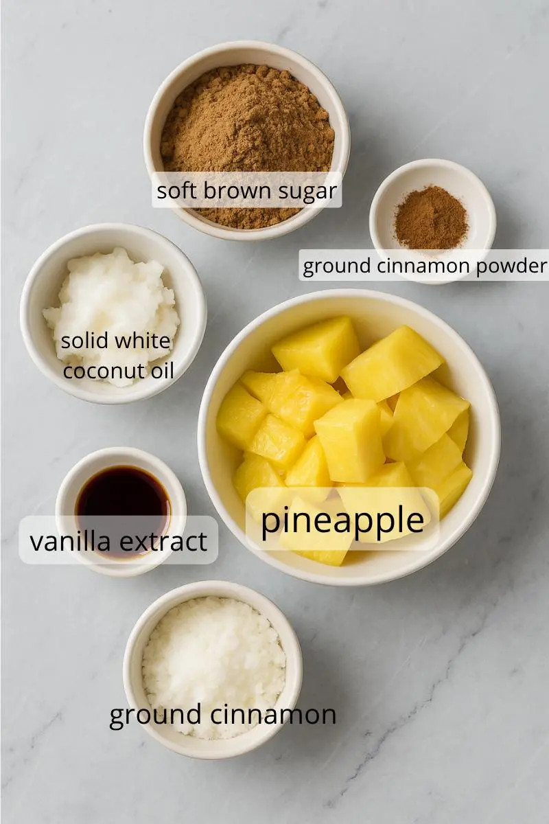 Overhead view of five cooking ingredients in white bowls on a grey marble countertop: pineapple chunks, coconut oil, brown sugar, ground cinnamon, and vanilla extract.